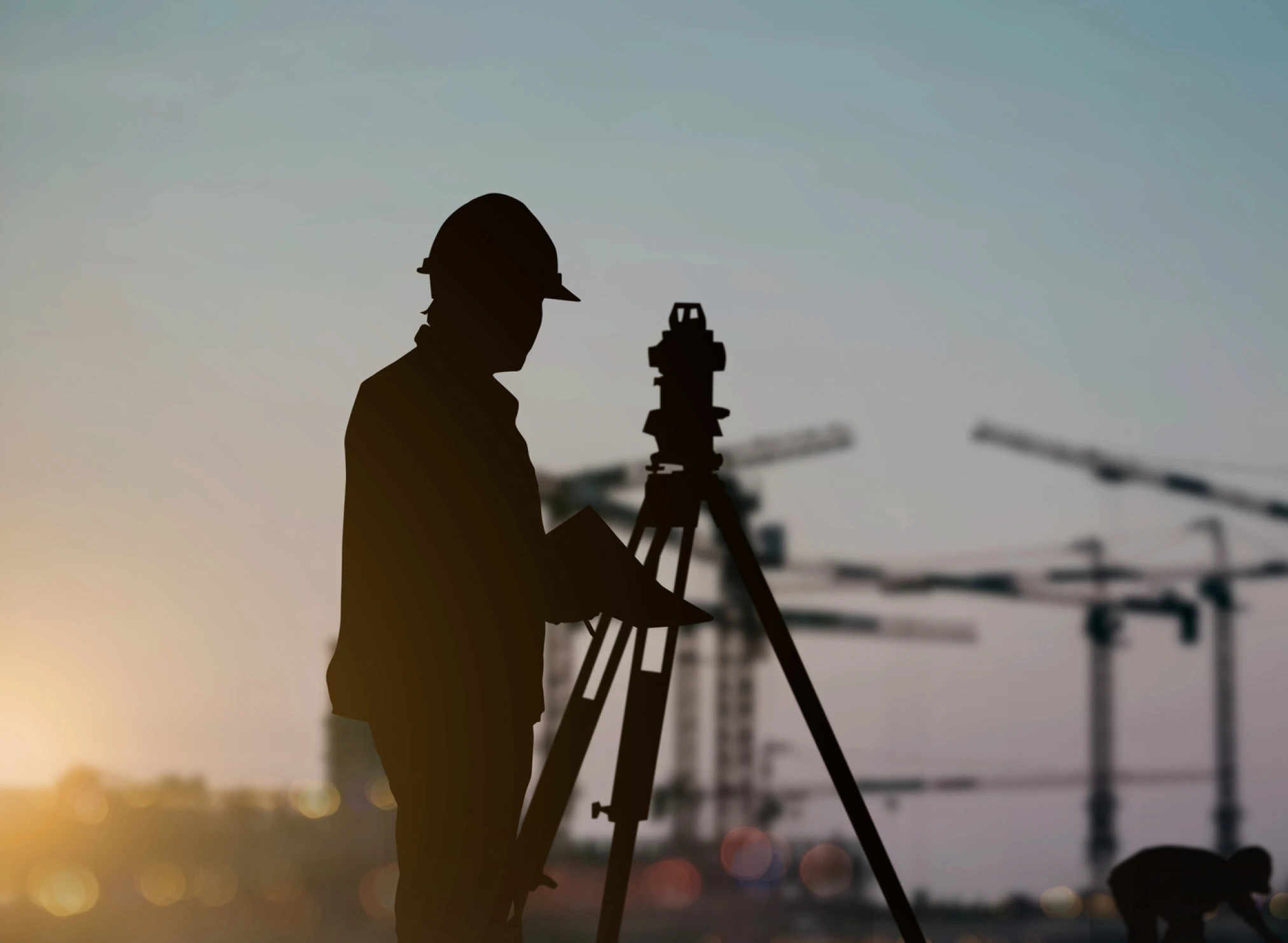The silhouette of a man in a hard hat and survey equipment with a build site off in the distance